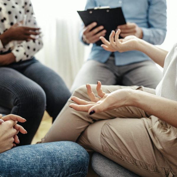 Unrecognizable vulnerable woman participates in group therapy session. Unrecognizable Caucasian person gestures while speaking. Group of people sitting in a circle during therapy.